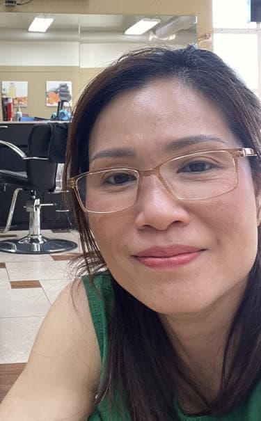 Smiling woman with brown hair and glasses wearing a green top in a hair salon.